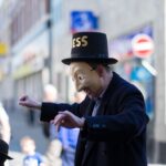 An actor in a mask wearing a top hat with the word 'Happiness' written on it